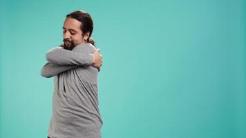 Portrait of man standing in studio, hugging himself in gesture of comfort and self love, isolated over studio background. Cheerful person doing self acceptance, embracing himself, camera B video