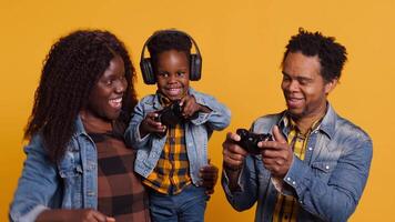 Enthusiastic father playing online games with baby boy using controller, both parents enjoying leisure time with their small kid. African american happy family play with joystick. Camera A. video