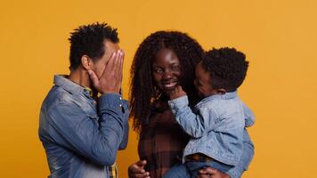 African american positive couple doing peek a boo with their young child in studio, smiling people hugging together. Happy affectionate mom and dad showing gratitude and respect. Camera A. video