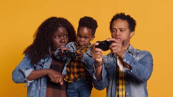 Cheerful african american dad playing games with his son on console, using controller to have fun against yellow background. Happy positive young family enjoying their leisure time. Camera A. video