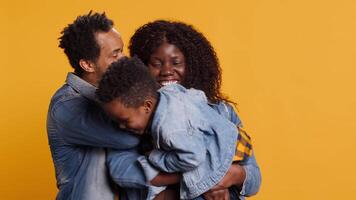 Joyful little child playing with a controller for games, posing with mother and father in studio. African american family with toddler standing against yellow background, kid gaming. Camera A. video