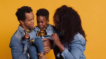 African american family and their little child in studio holding each other against yellow background. Smiling proud young couple posing with their adorable toddler, enjoying leisure time. Camera A. video