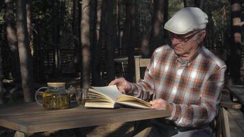 An elderly man sits at a wooden table, immersed in a book, surrounded by tall trees in a peaceful forest. He enjoys a moment of solitude with tea. video