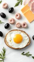 An egg, bread, cheese, olives and other foods are arranged on a white surface photo