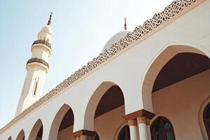 Low angle view of a white mosque with arches and minaret under a clear sky. photo