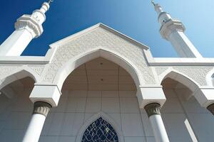 Low-angle view of a white mosque with intricate details, two minarets, and a clear blue sky. photo