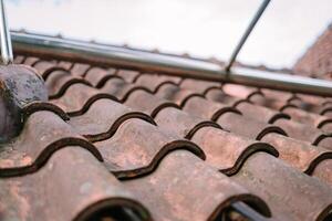 Close-up of rustic clay roof tiles in a wave pattern, with a metal pipe structure above. The weathered texture and mossy spots add character to the rooftop against a light sky. photo