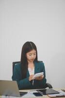 Asian businesswoman in red suit calculating company sales with calculator, laptop, and tablet on table Interior of businesswoman office at the modern workstation. photo