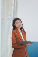Attractive smiling Asian businesswoman standing holding tablet working and recording work details in office. photo