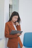 Attractive smiling Asian businesswoman standing holding tablet working and recording work details in office. photo