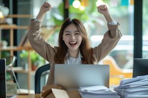 Happy excited young Korean business woman accountant standing at the desk working on laptop computer with a pile of documents on table in office and making yes gesture rejoicing in successful job. photo