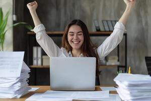 Happy excited young Hispanic business woman accountant standing at the desk working on laptop computer with a pile of documents on table in office and making yes gesture rejoicing in successful job. photo