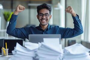 Happy excited young Indian business man accountant standing at the desk working on laptop computer with a pile of documents on table in office and making yes gesture rejoicing in successful job. photo