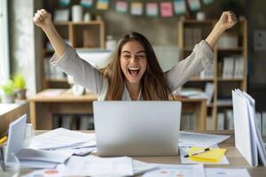 Happy excited young Hispanic business woman accountant standing at the desk working on laptop computer with a pile of documents on table in office and making yes gesture rejoicing in successful job. photo