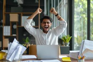 Happy excited young Indian business man accountant standing at the desk working on laptop computer with a pile of documents on table in office and making yes gesture rejoicing in successful job. photo