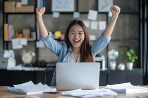 Happy excited young Korean business woman accountant standing at the desk working on laptop computer with a pile of documents on table in office and making yes gesture rejoicing in successful job. photo