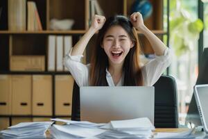 Happy excited young Korean business woman accountant standing at the desk working on laptop computer with a pile of documents on table in office and making yes gesture rejoicing in successful job. photo