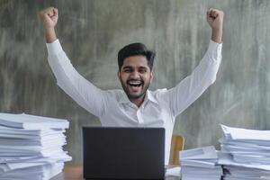 Happy excited young Indian business man accountant standing at the desk working on laptop computer with a pile of documents on table in office and making yes gesture rejoicing in successful job. photo