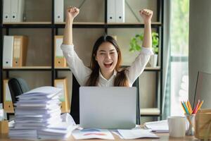 Happy excited young business woman accountant standing at the desk working on laptop computer with a pile of documents on table in office and making yes gesture rejoicing in successful job. photo