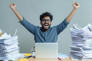 Happy excited young Indian business man accountant standing at the desk working on laptop computer with a pile of documents on table in office and making yes gesture rejoicing in successful job. photo