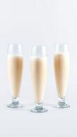 Three elegant glasses of creamy beverage stand in a row on a plain background, showcasing a light tan hue photo