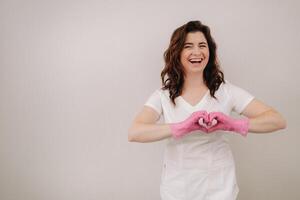 A smiling doctor in pink gloves and a white coat looks into the camera and makes a heart with his hands on a light background photo