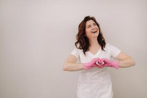 A smiling doctor in pink gloves and a white coat looks into the camera and makes a heart with his hands on a light background photo