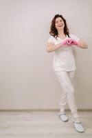 A smiling doctor in pink gloves and a white coat looks into the camera and makes a heart with his hands on a light background photo