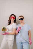 Two cosmetologists in protective glasses are holding a laser depilation device and a cactus in their hands photo