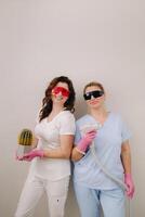 Two cosmetologists in protective glasses are holding a laser depilation device and a cactus in their hands photo