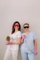Two cosmetologists in protective glasses are holding a laser depilation device and a cactus in their hands photo