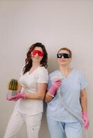 Two cosmetologists in protective glasses are holding a laser depilation device and a cactus in their hands photo
