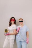 Two cosmetologists in protective glasses are holding a laser depilation device and a cactus in their hands photo