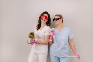 Two cosmetologists in protective glasses are holding a laser depilation device and a cactus in their hands photo