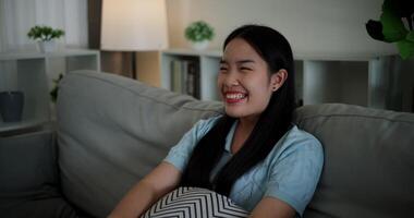 Handheld shot, Cheerful Asian woman watching something on TV and laughing happily while sitting on sofa in the living room at home. Leisure activity, Lifestyle concept. video