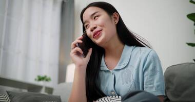 Handheld low angle view shot, Cheerful young woman is having fun talking on smartphone with her friend, smiling and laughing happily while sitting on the sofa in the living room at home. video