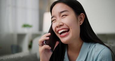 Handheld shot, Cheerful young woman is having fun talking on smartphone with her friend, smiling and laughing happily while sitting on the sofa in the living room at home. video