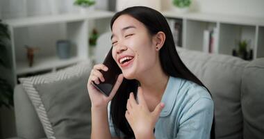 Handheld high angle view shot, Cheerful young woman is having fun talking on smartphone with her friend, smiling and laughing happily while sitting on the sofa in the living room at home. video
