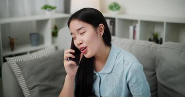 Handheld high angle view shot, Cheerful young woman is having fun talking on smartphone with her friend, smiling and laughing happily while sitting on the sofa in the living room at home. video