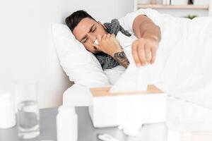 Cold, flue, coronavirus concept. Sick guy taking napkins from tissue box and sneezing runny nose, lying in bed with table full of pills nearby, copy space photo