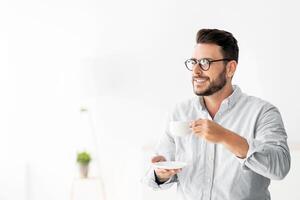 Young freelancer guy with cup of coffee standing at desk at home interior, leaning at table and looking away, enjoying hot morning drink, copy space photo