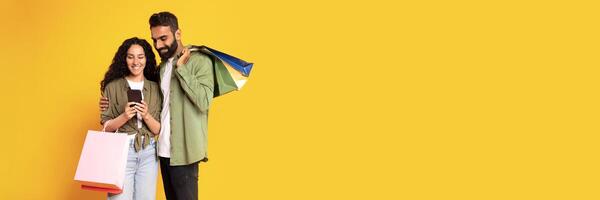 Mobile Shopping App. Happy Arab Couple Using Cellphone Purchasing Something Online In Application Standing With Colorful Shopper Bags On Yellow Background, Studio Shot. Ecommerce, Copy Space photo