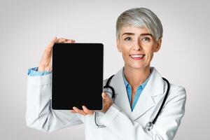 Smiling woman doctor showing tablet with blank screen, isolated on light background, studio shot photo