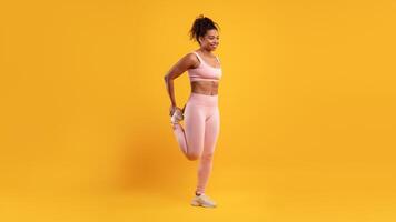 A young woman in pink activewear performs a leg stretch before starting her exercise routine in a vibrant yellow studio. Her focused expression shows dedication to fitness. photo