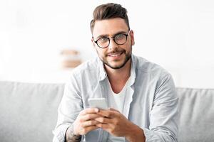 Positive young guy with glasses sitting on couch, using smartphone and smiling to camera, chatting with friends and chilling at home, copy space photo