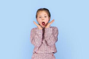 A joyful girl with a surprised expression raises her hands to her face, showcasing her happiness. She is dressed in a stylish outfit, against a simple blue backdrop, highlighting her playful emotions. photo