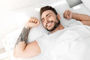 Happy wellslept guy waking up in morning, stretching while lying in comfortable bed in bedroom at home, smiling and looking at window, high-angle view. Good morning, awakening concept. photo