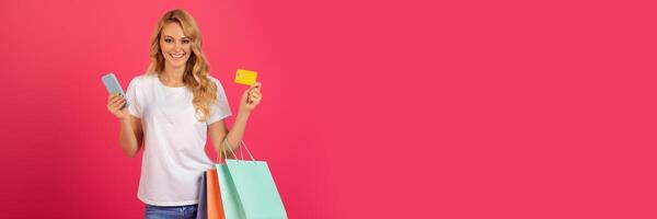 Online Payment. Happy blonde lady shopping holding credit card and mobile phone, ordering clothing purchase on internet, posing with paper shopper bags over bright pink studio backdrop, copy space photo