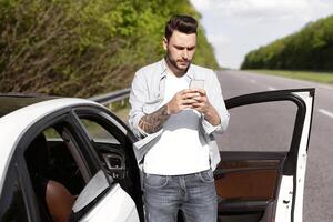 Upset young man standing near car, looking at smartphone screen, having problem with mobile connection, trying to find network on highway. Road emergencies, calling breakdown service, no signal photo