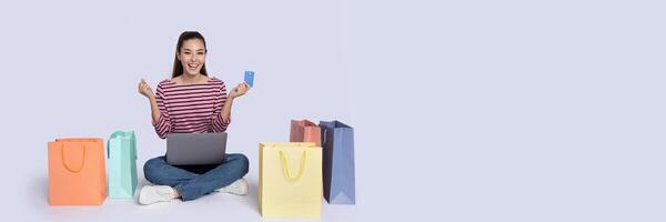 A happy shopper sits on the floor surrounded by colorful shopping bags while holding a credit card and a laptop. She enjoys the thrill of online shopping for great Black Friday deals, copy space photo